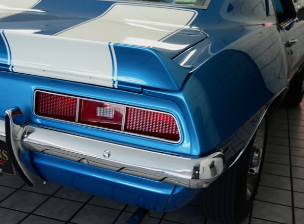 A close-up view of the rear section of a classic car, featuring a blue paint job with white racing stripes. The car's chrome bumper and taillights are prominently visible, showcasing a vintage design.