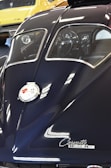 A detailed view of the rear of a classic Corvette Sting Ray, showcasing its sleek, dark blue body and split rear window design. The chrome emblem and cursive 'Corvette Sting Ray' label are prominently visible. Reflections on the car's surface suggest bright overhead lighting. In the background, a yellow classic car is partially visible.