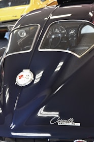 A detailed view of the rear of a classic Corvette Sting Ray, showcasing its sleek, dark blue body and split rear window design. The chrome emblem and cursive 'Corvette Sting Ray' label are prominently visible. Reflections on the car's surface suggest bright overhead lighting. In the background, a yellow classic car is partially visible.