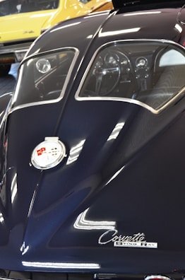 A detailed view of the rear of a classic Corvette Sting Ray, showcasing its sleek, dark blue body and split rear window design. The chrome emblem and cursive 'Corvette Sting Ray' label are prominently visible. Reflections on the car's surface suggest bright overhead lighting. In the background, a yellow classic car is partially visible.