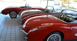 A showroom with a row of vintage red convertible cars lined up side by side on a tiled floor. The cars feature a classic design with chrome accents and whitewall tires. The interior of the cars is visible, showcasing dashboards and steering wheels. Sunlight streams in through the large glass windows, enhancing the shiny surfaces of the vehicles.