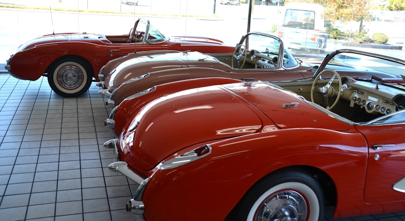 A showroom with a row of vintage red convertible cars lined up side by side on a tiled floor. The cars feature a classic design with chrome accents and whitewall tires. The interior of the cars is visible, showcasing dashboards and steering wheels. Sunlight streams in through the large glass windows, enhancing the shiny surfaces of the vehicles.