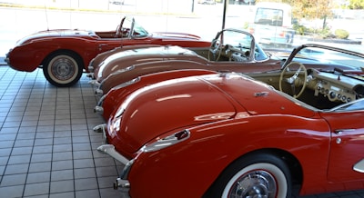A showroom with a row of vintage red convertible cars lined up side by side on a tiled floor. The cars feature a classic design with chrome accents and whitewall tires. The interior of the cars is visible, showcasing dashboards and steering wheels. Sunlight streams in through the large glass windows, enhancing the shiny surfaces of the vehicles.