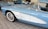 A close-up view of a vintage light blue and white car with a prominent front fender and a checkered flag emblem. The car is parked indoors on a tiled floor with other classic cars visible in the background. The vehicle has white-walled tires and shiny chrome side mirrors.