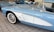 A close-up view of a vintage light blue and white car with a prominent front fender and a checkered flag emblem. The car is parked indoors on a tiled floor with other classic cars visible in the background. The vehicle has white-walled tires and shiny chrome side mirrors.