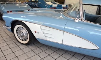 A close-up view of a vintage light blue and white car with a prominent front fender and a checkered flag emblem. The car is parked indoors on a tiled floor with other classic cars visible in the background. The vehicle has white-walled tires and shiny chrome side mirrors.