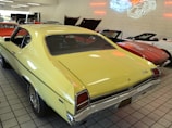 A collection of vintage cars is showcased in a showroom with tile flooring. The prominent car is a yellow classic model with a black stripe on the trunk. Other cars, including a red convertible, are visible in the background. Neon outlines of cars are displayed on the wall, adding a retro feel.