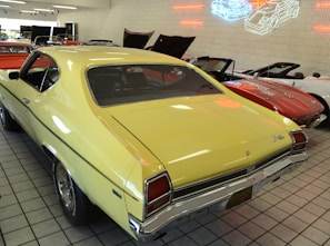 A collection of vintage cars is showcased in a showroom with tile flooring. The prominent car is a yellow classic model with a black stripe on the trunk. Other cars, including a red convertible, are visible in the background. Neon outlines of cars are displayed on the wall, adding a retro feel.