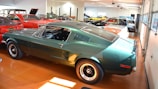 Various classic cars are displayed in a well-lit showroom with a predominance of a green vintage Mustang in the foreground. The polished floor complements the shiny exteriors of the vehicles, creating a sense of nostalgia and elegance.