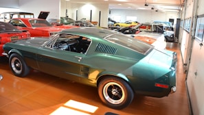 Various classic cars are displayed in a well-lit showroom with a predominance of a green vintage Mustang in the foreground. The polished floor complements the shiny exteriors of the vehicles, creating a sense of nostalgia and elegance.