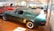 Various classic cars are displayed in a well-lit showroom with a predominance of a green vintage Mustang in the foreground. The polished floor complements the shiny exteriors of the vehicles, creating a sense of nostalgia and elegance.