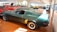Various classic cars are displayed in a well-lit showroom with a predominance of a green vintage Mustang in the foreground. The polished floor complements the shiny exteriors of the vehicles, creating a sense of nostalgia and elegance.