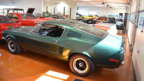 Various classic cars are displayed in a well-lit showroom with a predominance of a green vintage Mustang in the foreground. The polished floor complements the shiny exteriors of the vehicles, creating a sense of nostalgia and elegance.