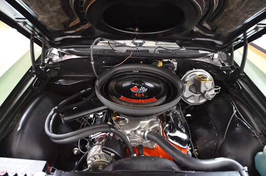 A close-up view of a car engine inside the hood, showcasing a powerful 454 cubic inch V8 engine. The air cleaner has a checkered flag decal and the label 'Turbo Jet 450 HP'. Various metallic and rubber components are visible, with the engine block painted in orange.