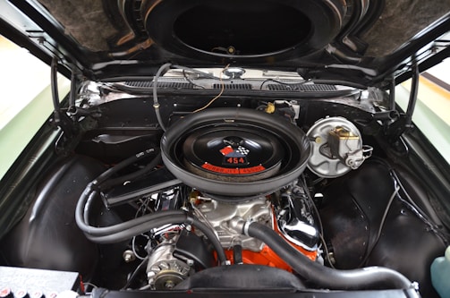 A close-up view of a car engine inside the hood, showcasing a powerful 454 cubic inch V8 engine. The air cleaner has a checkered flag decal and the label 'Turbo Jet 450 HP'. Various metallic and rubber components are visible, with the engine block painted in orange.