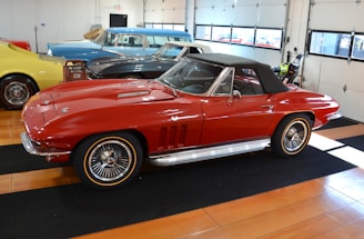 A classic red convertible car is parked inside a well-lit garage with a wooden floor. The car features sleek lines, chrome accents, and a black soft top. In the background, other vintage cars in blue, black, and yellow can be seen, suggesting a collection or showroom setting.
