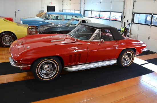 A classic red convertible car is parked inside a well-lit garage with a wooden floor. The car features sleek lines, chrome accents, and a black soft top. In the background, other vintage cars in blue, black, and yellow can be seen, suggesting a collection or showroom setting.