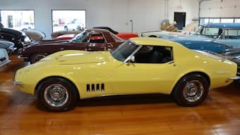 A collection of classic cars displayed indoors, prominently featuring a yellow sports car with sleek curves and shiny chrome wheels. The cars are positioned on a polished wooden floor under bright lighting. Large windows in the background allow natural light to illuminate the showroom, and other vintage vehicles in various colors are lined up behind and beside the car.