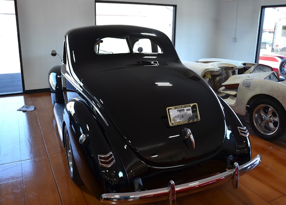 A vintage black car is parked indoors on a wooden floor. The car has a glossy finish with reflections of ceiling lights. Its rear license plate displays an Ohio registration. Surrounding the car are additional vehicles being worked on, with windows letting in natural light from outside.