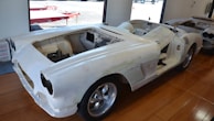 A partially assembled classic car frame sits on a wooden floor inside a garage or workshop. The body is unpainted, with visible primer, and it lacks doors, a hood, and other components. Large, shiny wheels are mounted, and parts of the interior are exposed.