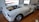 A partially assembled classic car frame sits on a wooden floor inside a garage or workshop. The body is unpainted, with visible primer, and it lacks doors, a hood, and other components. Large, shiny wheels are mounted, and parts of the interior are exposed.