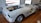A partially assembled classic car frame sits on a wooden floor inside a garage or workshop. The body is unpainted, with visible primer, and it lacks doors, a hood, and other components. Large, shiny wheels are mounted, and parts of the interior are exposed.