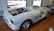 A partially assembled classic car frame sits on a wooden floor inside a garage or workshop. The body is unpainted, with visible primer, and it lacks doors, a hood, and other components. Large, shiny wheels are mounted, and parts of the interior are exposed.