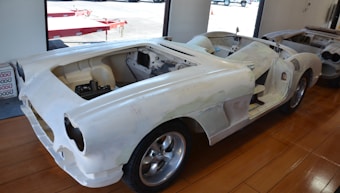 A partially assembled classic car frame sits on a wooden floor inside a garage or workshop. The body is unpainted, with visible primer, and it lacks doors, a hood, and other components. Large, shiny wheels are mounted, and parts of the interior are exposed.