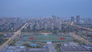 A sprawling cityscape featuring numerous high-rise buildings under a blue, overcast sky. In the foreground, an athletics track and field are visible, surrounded by lush greenery and organized urban planning. Streets are well-lit with streetlights, and traffic flows smoothly along the roads. The image captures the blend of nature with urban development, highlighting both buildings and recreational spaces.