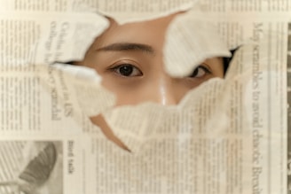 A person's eyes peer through a torn hole in a newspaper, revealing a close-up view of one of their eyes and part of their face. The newspaper text is visible around the torn edges.