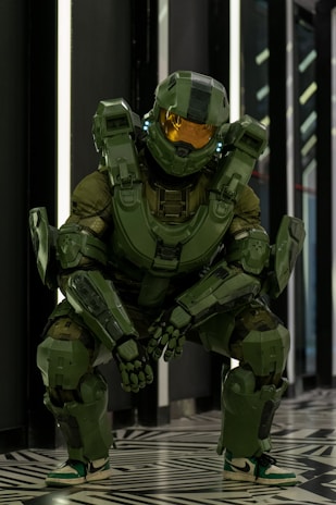 A person wearing a detailed suit of futuristic green armor stands in an interior space with patterned flooring. The armor is robust, featuring angular plates, a helmet with a reflective visor, and illuminated accents. The setting is modern, with dark walls and bright vertical lights.