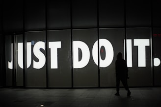 A large, illuminated sign with bold white letters spelling out 'JUST DO IT.' stands prominently against a dark background. In the foreground, a silhouette of a person in a hoodie is visible, facing the sign, adding a sense of intrigue and mystery.