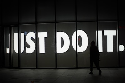 A large, illuminated sign with bold white letters spelling out 'JUST DO IT.' stands prominently against a dark background. In the foreground, a silhouette of a person in a hoodie is visible, facing the sign, adding a sense of intrigue and mystery.