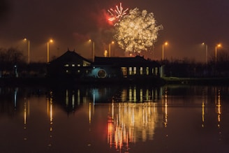 Fireworks burst in a night sky above a building, with reflections visible on a calm body of water. Streetlights line the background, casting a warm glow.