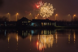 Fireworks burst in a night sky above a building, with reflections visible on a calm body of water. Streetlights line the background, casting a warm glow.