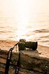 A digital camera with a zoom lens and branded strap rests on a stone ledge overlooking a body of water. The sun reflects off the water, creating a warm, golden glow.