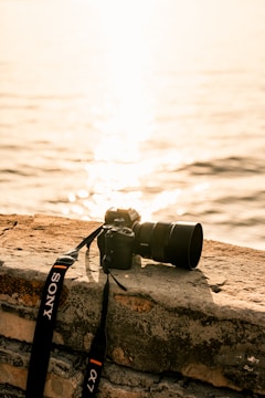 A digital camera with a zoom lens and branded strap rests on a stone ledge overlooking a body of water. The sun reflects off the water, creating a warm, golden glow.