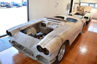 Two vintage car bodies are placed in a workshop with a wooden floor and storage area. Both cars are missing engines and other components, indicating they are in the process of restoration. The interior space is bright due to large windows, allowing ample natural light to illuminate the scene.