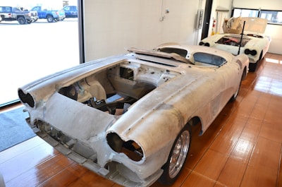 Two vintage car bodies are placed in a workshop with a wooden floor and storage area. Both cars are missing engines and other components, indicating they are in the process of restoration. The interior space is bright due to large windows, allowing ample natural light to illuminate the scene.