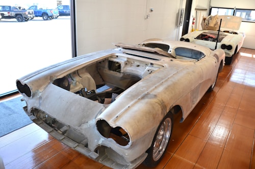 Two vintage car bodies are placed in a workshop with a wooden floor and storage area. Both cars are missing engines and other components, indicating they are in the process of restoration. The interior space is bright due to large windows, allowing ample natural light to illuminate the scene.