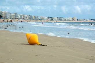 A sandy beach with a yellow buoy lying on the shore, surrounded by gentle waves. In the background, modern apartment buildings line the coast, and numerous people are enjoying activities along the water and on the beach.