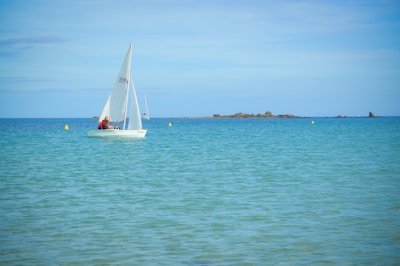 A small white sailboat with several people on board glides across calm turquoise waters. The sea stretches into the distance, meeting a clear blue sky. A few yellow buoys float nearby, and a rocky landmass is visible on the horizon.