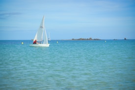A small white sailboat with several people on board glides across calm turquoise waters. The sea stretches into the distance, meeting a clear blue sky. A few yellow buoys float nearby, and a rocky landmass is visible on the horizon.