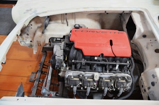 A partially disassembled car with a visible engine compartment. The centerpiece is a red engine cover with the word 'Corvette' and '5.7L' prominently displayed, indicating a V8 engine. The engine is installed within a chassis with a bare metal and unfinished body frame surrounding it.