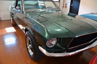 A classic green car with polished chrome accents is parked indoors on a wooden floor. The car's shiny surface reflects the surroundings, and the details like the headlights, tires, and front grille are clearly visible.