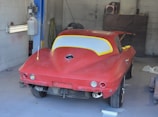 A partially restored or prepared vintage red car is inside a garage, with masking paper covering the windows and some unfinished or missing parts. The setting includes a concrete wall and typical garage equipment.