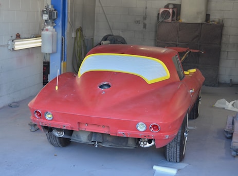 A partially restored or prepared vintage red car is inside a garage, with masking paper covering the windows and some unfinished or missing parts. The setting includes a concrete wall and typical garage equipment.