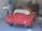 A partially restored or prepared vintage red car is inside a garage, with masking paper covering the windows and some unfinished or missing parts. The setting includes a concrete wall and typical garage equipment.