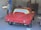 A partially restored or prepared vintage red car is inside a garage, with masking paper covering the windows and some unfinished or missing parts. The setting includes a concrete wall and typical garage equipment.
