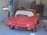 A partially restored or prepared vintage red car is inside a garage, with masking paper covering the windows and some unfinished or missing parts. The setting includes a concrete wall and typical garage equipment.